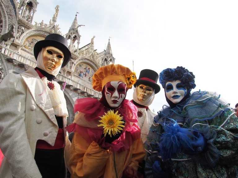 Karneval v Benátkách – čtyři postavy v barevných maskách a kostýmech před bazilikou sv. Marka